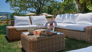 Zona relax esterna con mobili in vimini, un divano bianco, cuscini e un tavolino in vetro con snack e bevande.