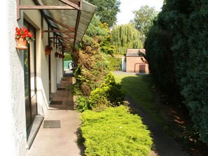 A pathway leading to a building with a covered porch, surrounded by greenery and a hedge.