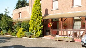 A building with a brick facade, a covered porch, and a tall coniferous tree in front. - Résidence Rachel 23 - Shape - Mons (Mons)