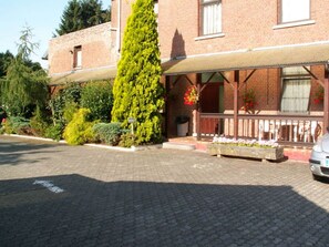 A building with a brick facade, a covered porch, and a tall coniferous tree in front. - Résidence Rachel 23 - Shape - Mons (Mons)