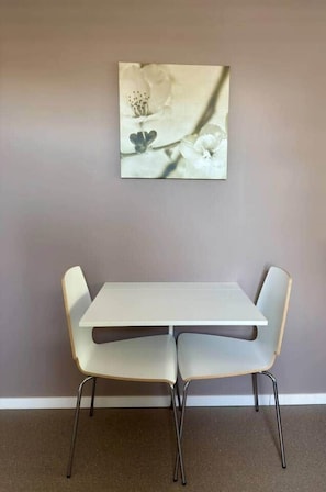 A dining area with a white table and two chairs, a wall-mounted artwork, and a carpeted floor. - Résidence Rachel 29 - Shape - Mons (Mons)
