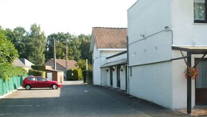 A quiet street with a red car parked on the side, a white building with a tiled roof, and a flower arrangement near the entrance.