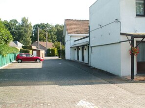 Une rue tranquille où est garée une voiture rouge, un bâtiment blanc avec un toit en tuiles et un arrangement floral près de l’entrée.