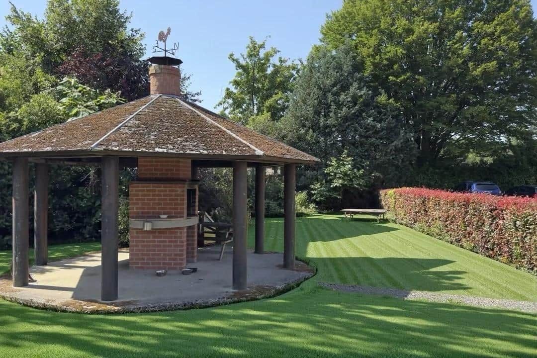 Ein Pavillon mit Backsteinfundament und einem Ziegeldach, umgeben von einem gepflegten Rasen und Hecken.