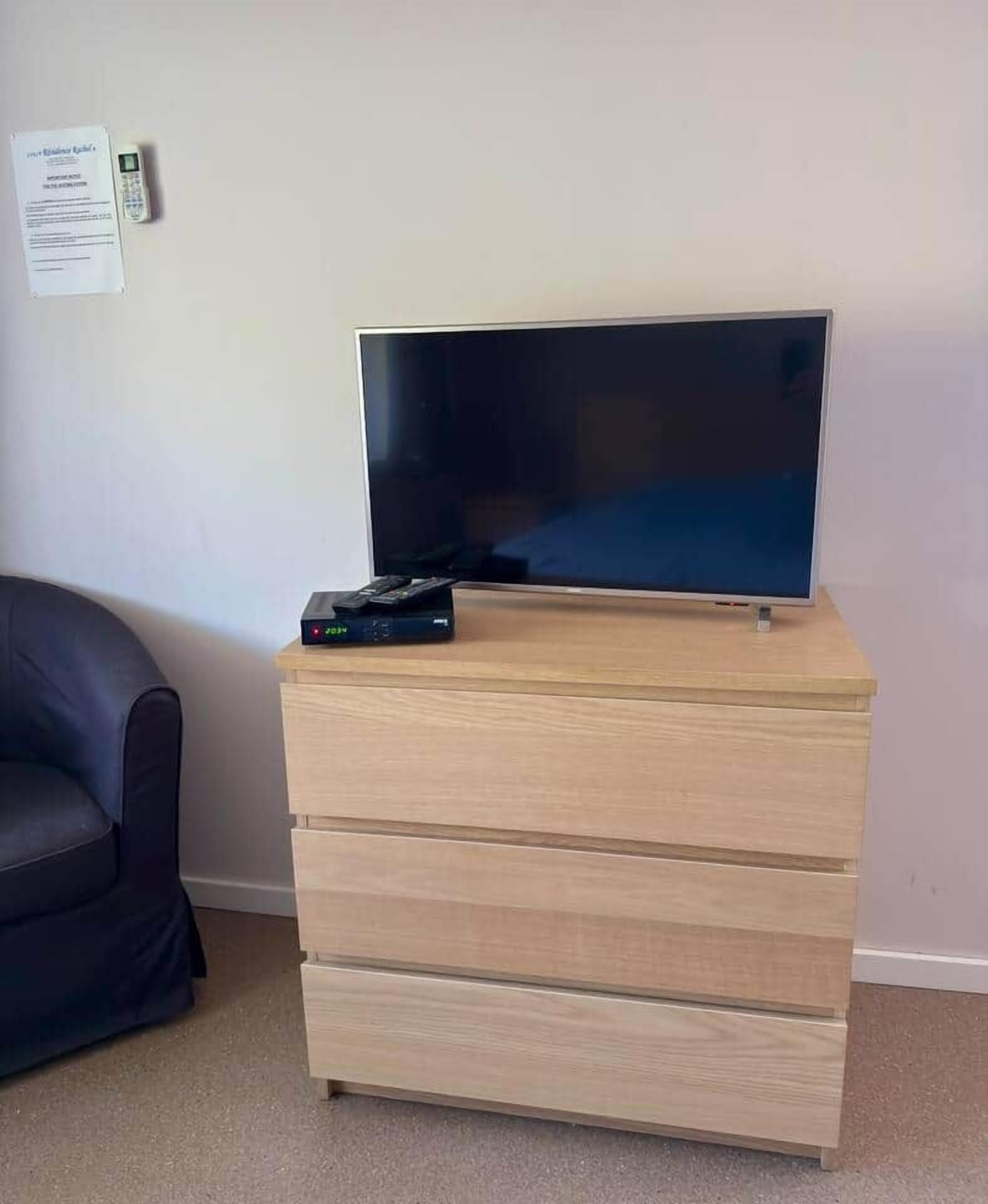 Ein Zimmer mit einem Flachbildfernseher auf einer Holzkommode, einer Fernbedienung und einem Dokument an der Wand.