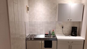 Een kleine keuken met een kookplaat, gootsteen en kastjes, een koffiezetapparaat, en een klein rood voorwerp op het aanrecht.