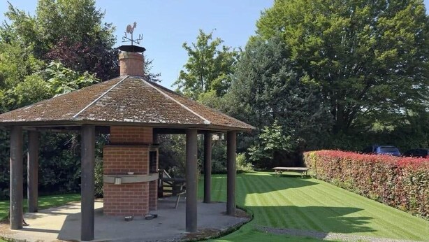 Een tuinpaviljoen met een fundering van baksteen en een pannendak, omringd door een verzorgd gazon en hagen.