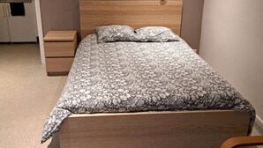 Een slaapkamer met een houten bed, nachtkastje, klein houten lade, wandlamp en een schilderij.