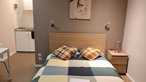 Una camera da letto con un letto, un comodino, una lampada a parete, un quadro e, sullo sfondo, un angolo cottura.