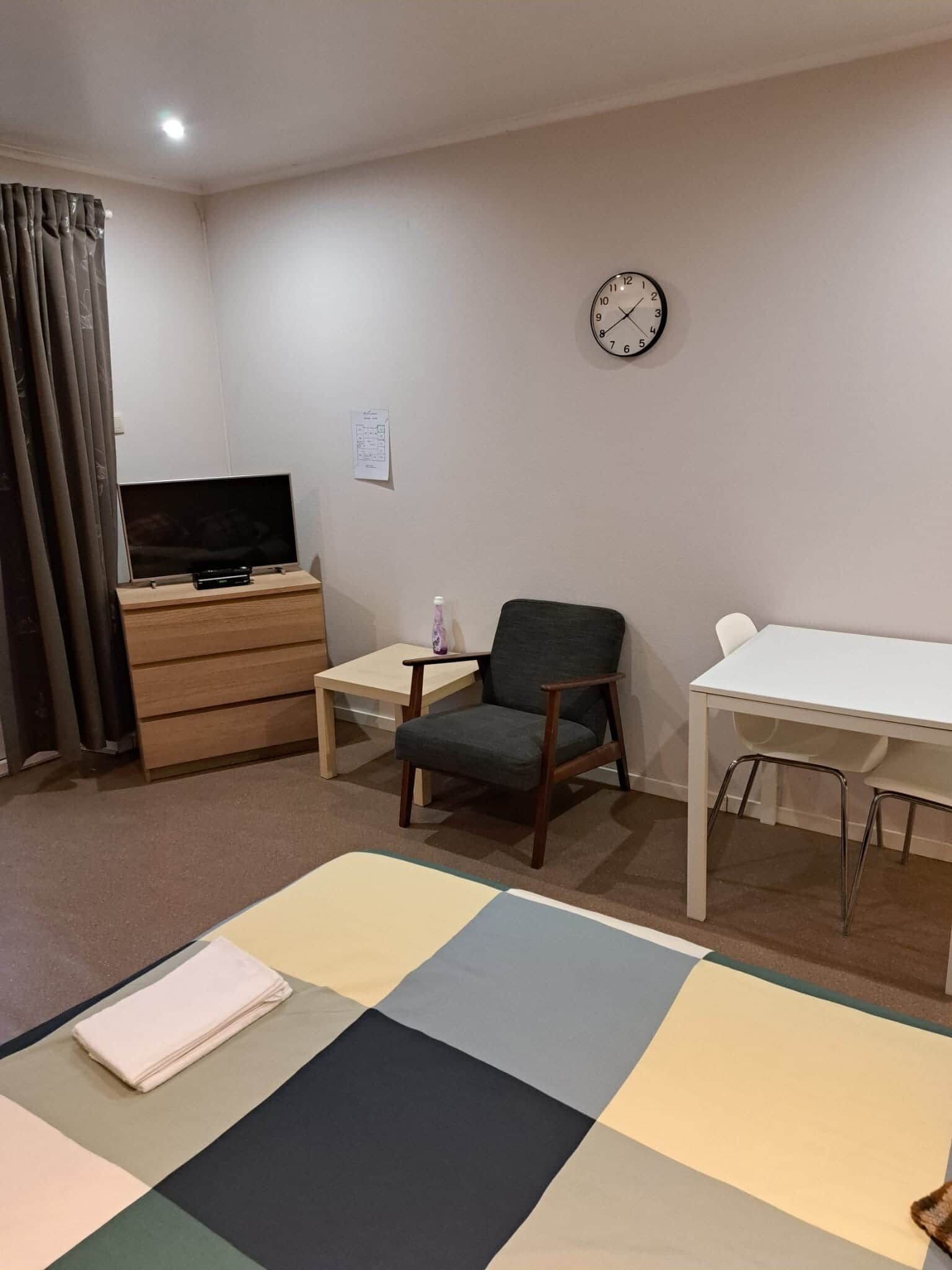 A hotel room with a bed, a TV on a wooden dresser, a chair, a small table, and a clock on the wall.