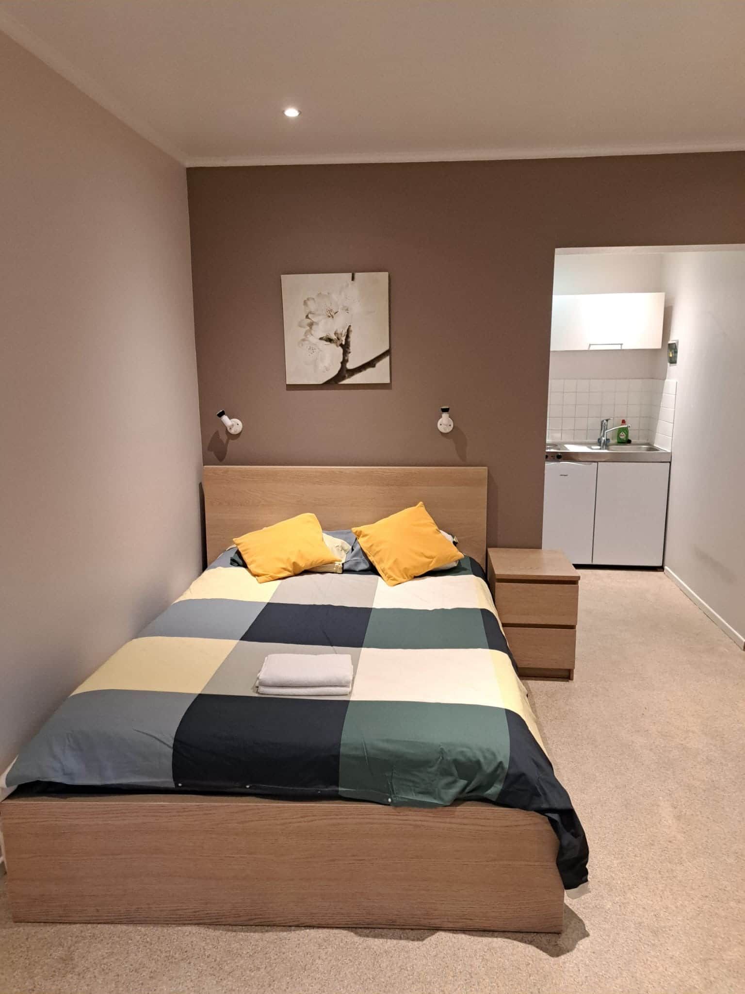 Une chambre à coucher comprenant un lit, des tables de chevet, une commode, une lampe fixée au mur, un tableau et un coin cuisine.