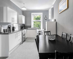 A modern kitchen with white cabinets, a dining table, and a window with a view of greenery. - Breathtaking Sea-View Apartment Steps from Rothesay Harbor (Rothesay)