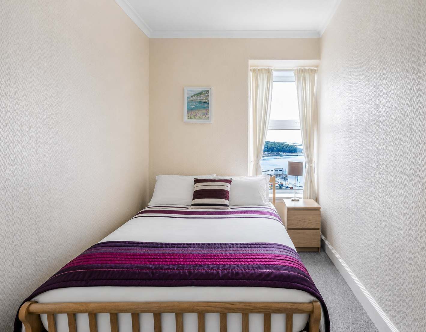 A narrow hotel room with a bed, a nightstand, a lamp, and a window with a view of the sea.