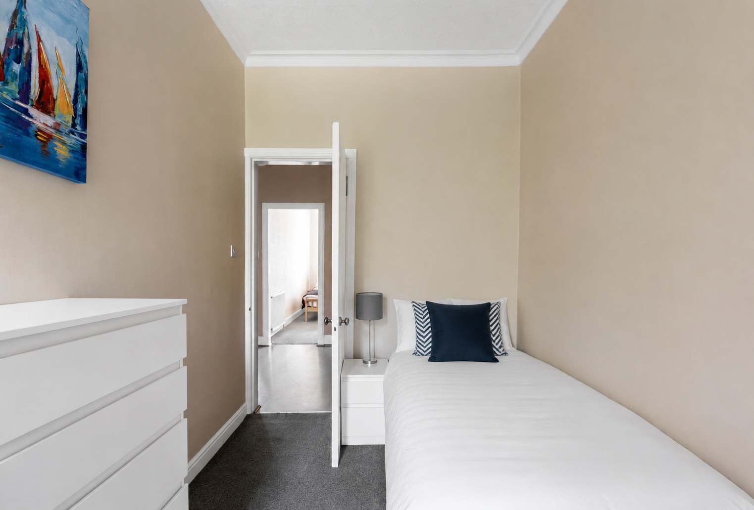 Una camera d'albergo con un letto, un comodino con una lampada, una cassettiera e un quadro appeso al muro.