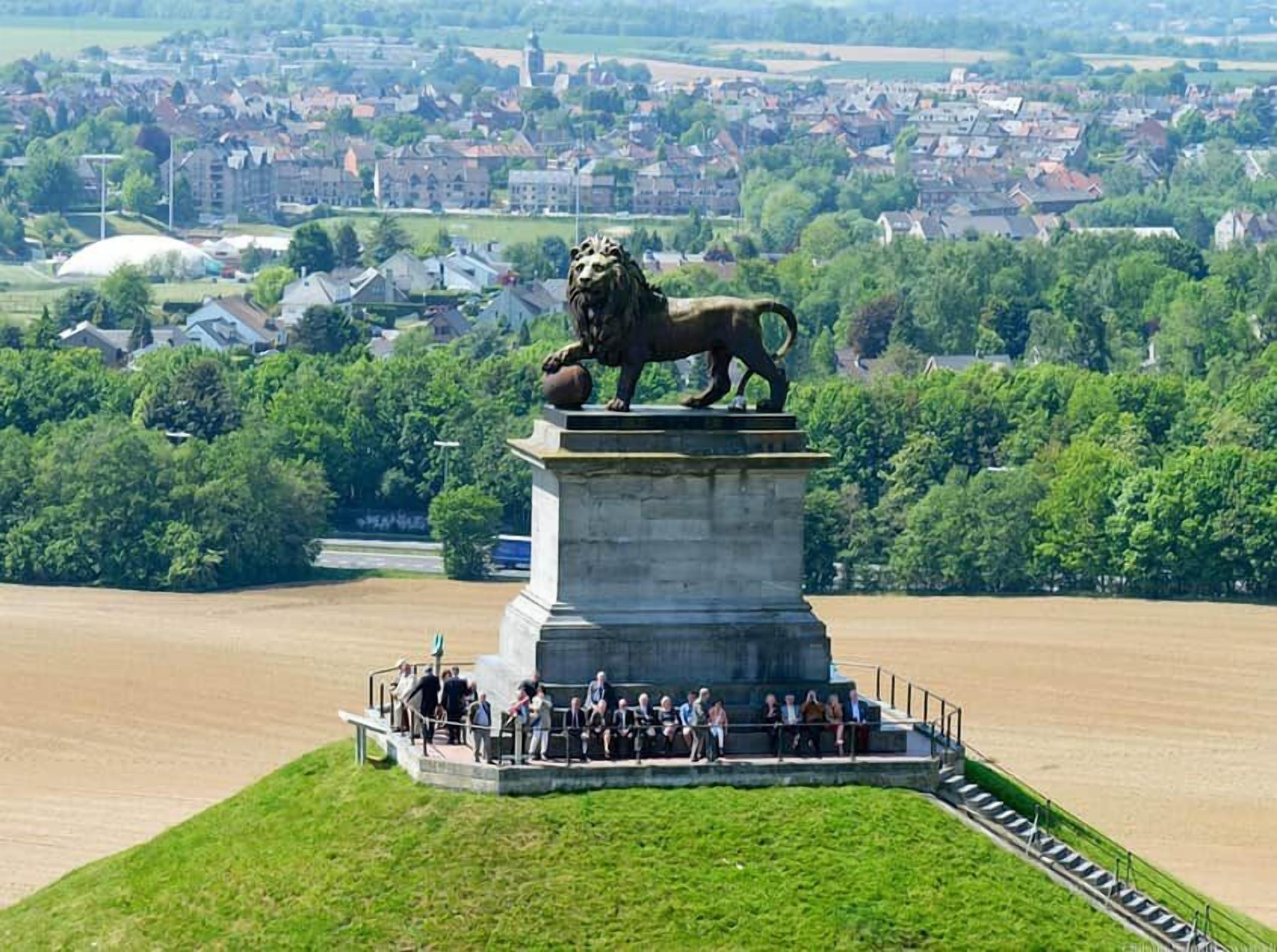 Une statue de lion, posée sur un piédestal, entourée d’un groupe de personnes, avec en arrière-plan une ville et une rivière.