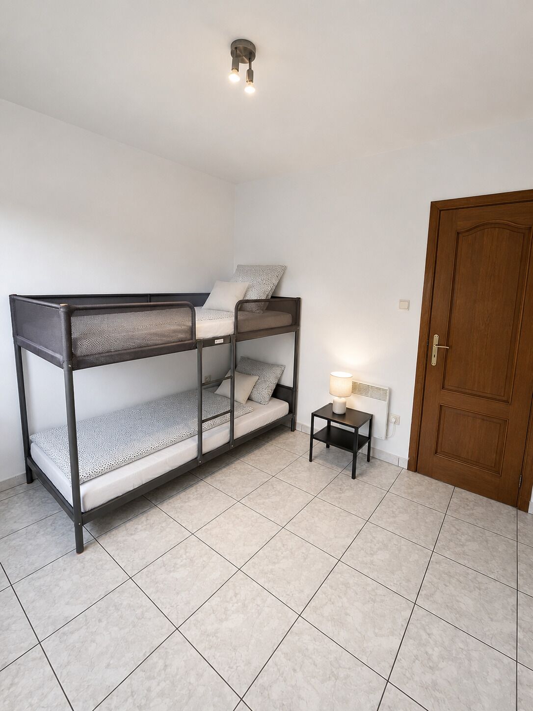 Una habitación con una cama litera, una mesita de noche pequeña con una lámpara, una puerta y una lámpara en el techo.
