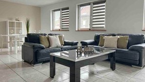 Una sala de estar con dos sofás, una mesa de centro, una mesa de comedor con sillas y dos ventanas con persianas.