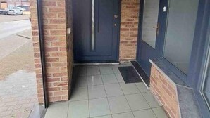Una entrada de edificio con una puerta oscura, un porche con baldosas y un muro de ladrillo.