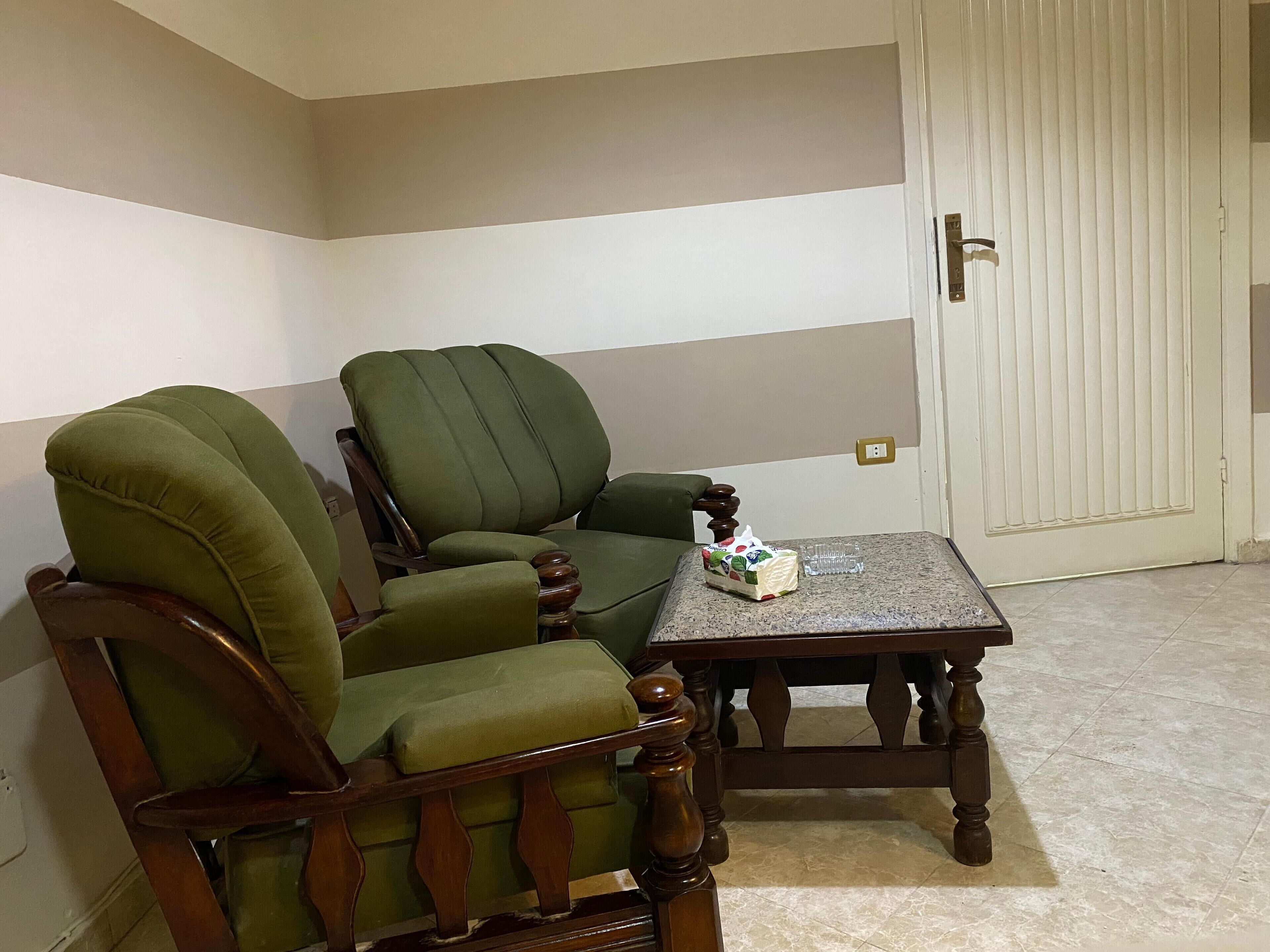 A corner room with a green armchair and a wooden table, a door, and a light switch on the wall.