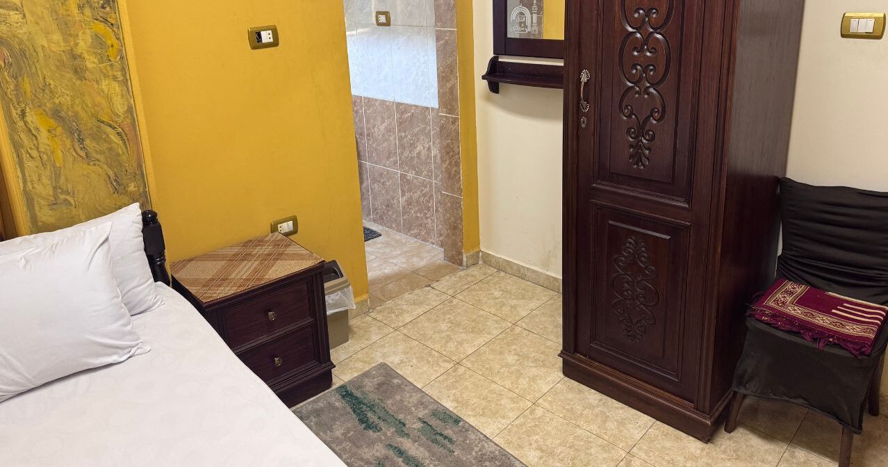 A hotel room with a bed, a wooden nightstand, a wardrobe, and a bathroom visible through an open door.