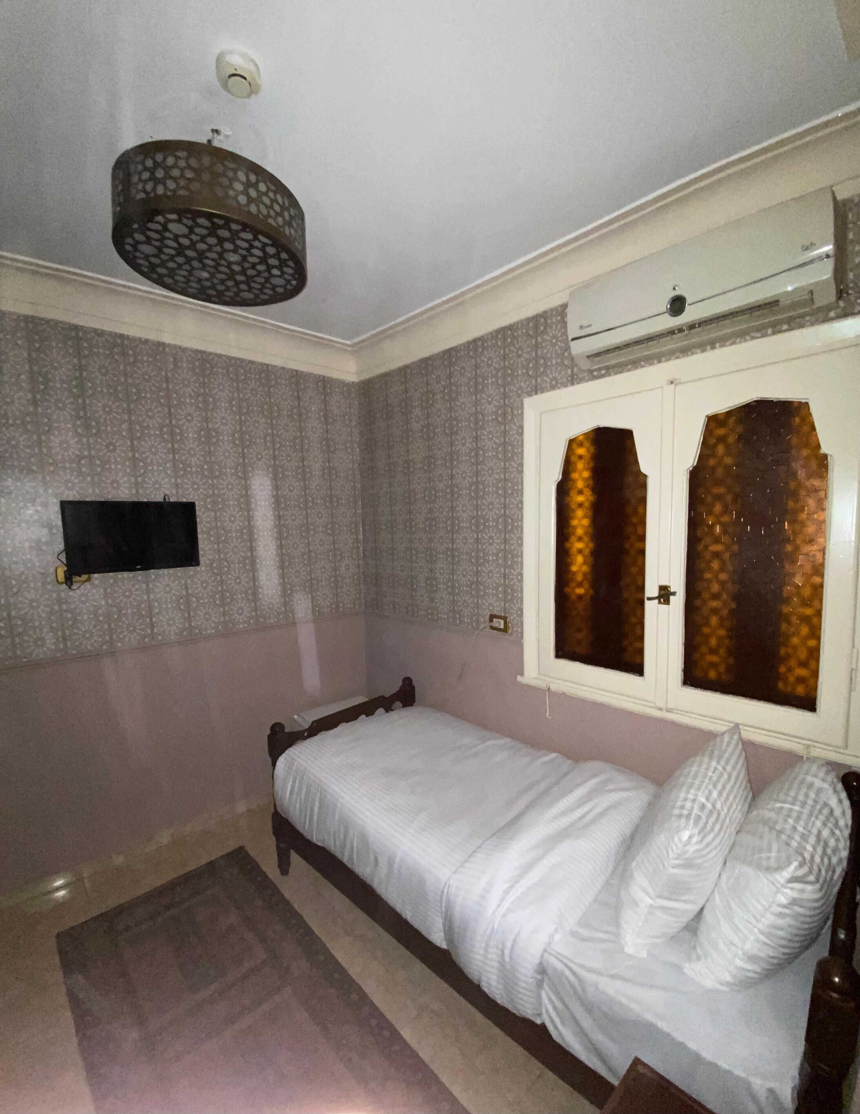 Una habitación pequeña con cama individual, papel tapiz estampado, televisor montado y ventana con vidrios decorativos.