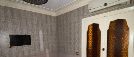 Una habitación pequeña con cama individual, papel tapiz estampado, televisor montado y ventana con vidrios decorativos.