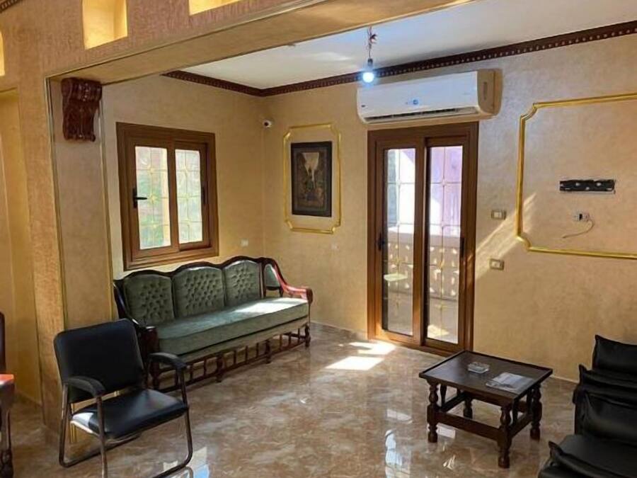 A spacious living area with a green sofa, a wooden coffee table, and a chair. There is a large window providing natural light and a door leading to an outdoor balcony.