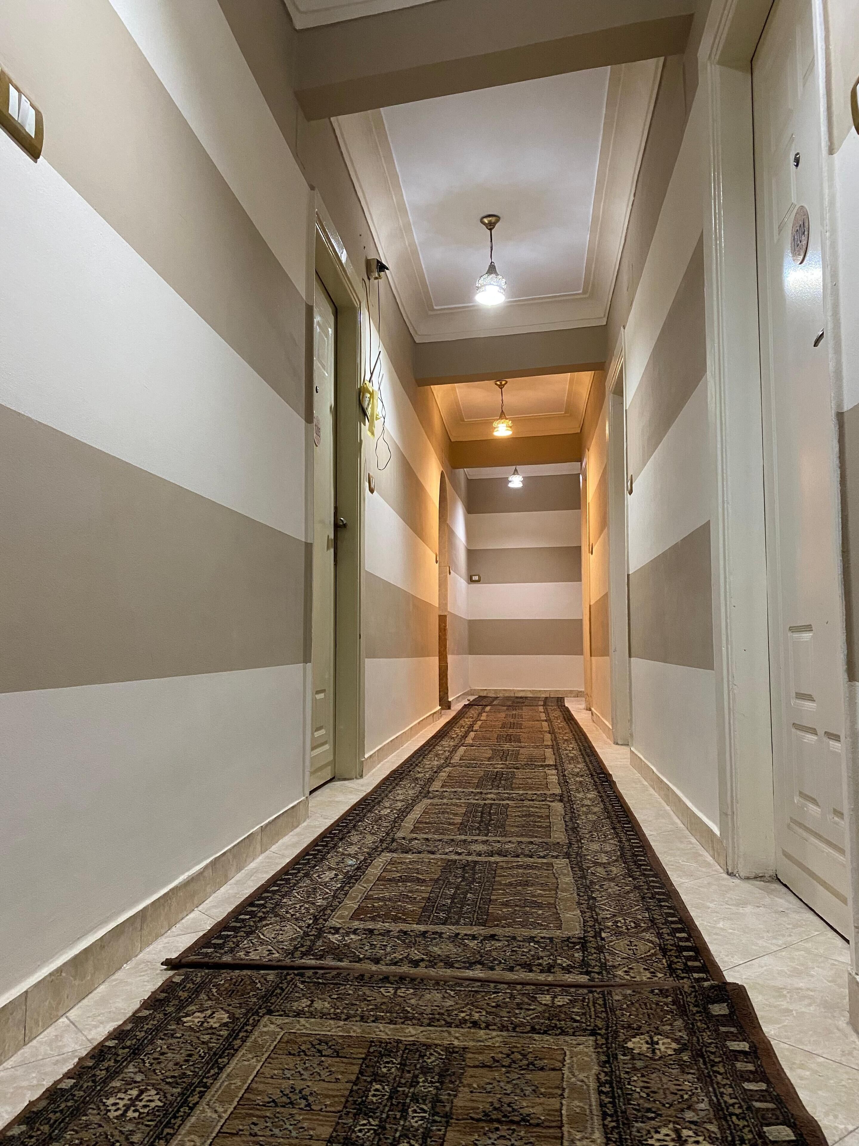 Un pasillo con alfombra estampada, paredes blancas con rayas horizontales y un ventilador de techo.