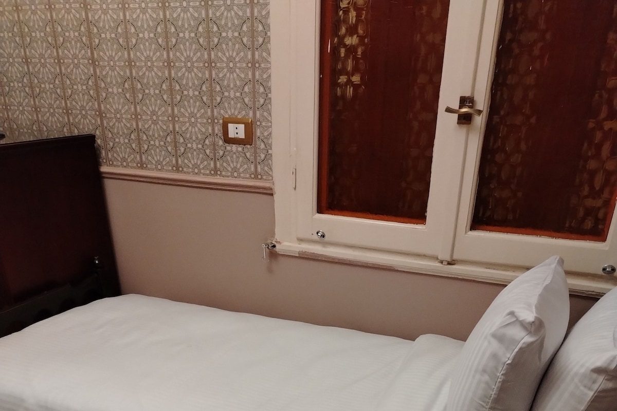 Quarto individual com uma cama, um tapete estampado, uma mesa de cabeceira de madeira e uma janela com vista para uma porta.