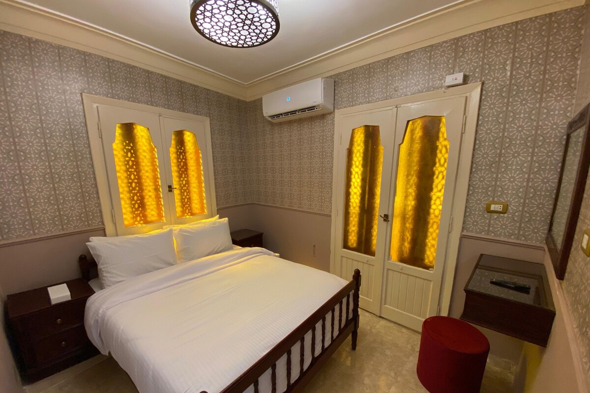 A bedroom with a bed, nightstands, a red stool, a desk, and a wardrobe with decorative glass panels.
