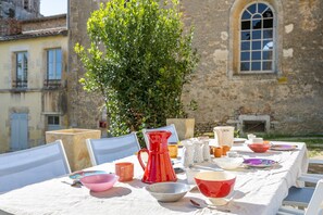 A table set outdoors with various dishes and a pitcher, surrounded by chairs, with a stone building and greenery in the background.