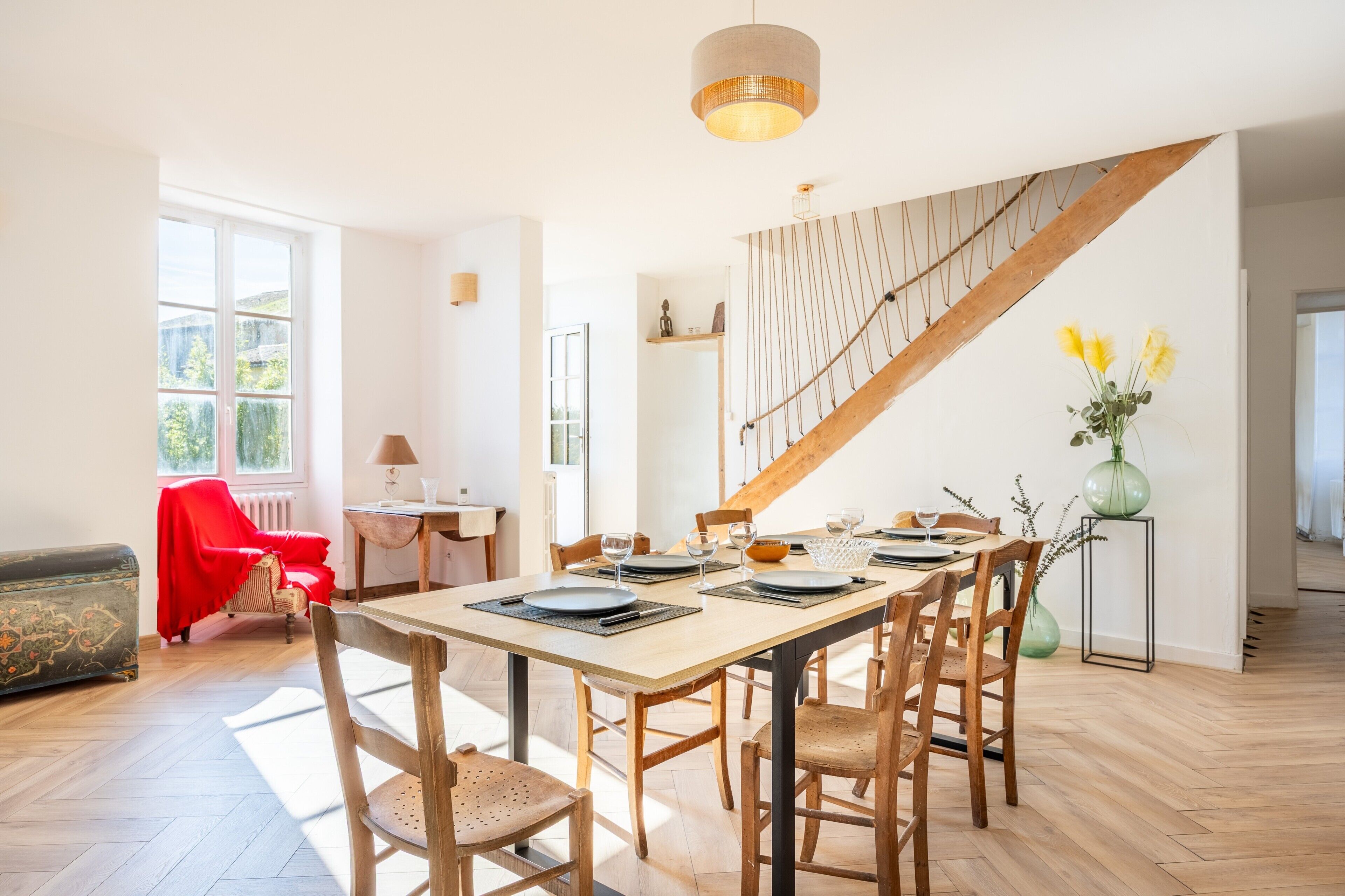 Un coin repas avec une table en bois dressée pour six personnes, un escalier et une vue sur la verdure à l’extérieur.