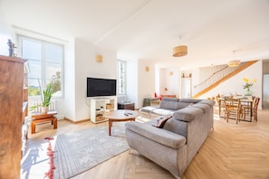 A spacious living room with a large sofa, a TV, and a dining area with a staircase. - Gîte du Val d’Egray – Charming House with Garden in Champdeniers (Champdeniers)