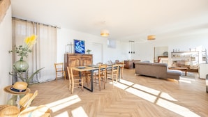 Amplia sala de estar con piso de madera en dibujo de espiga, una mesa de comedor con sillas, un sofá y una zona de cocina al fondo.