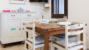 A dining area with a wooden table, white chairs, a white cabinet with drawers, a television mounted on the wall, and a window with a view. - GuestHost - Cozy detached house of about 60 sqm, perfect for 4 people, located on the ground floor.The property is located in Torre dei Corsari, a charming village overlooking the splendid Costa Verde of Sardinia. In a few minutes by car you can reach so (Torre dei Corsari)