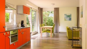 Une cuisine moderne avec des meubles de couleur orange, un coin repas avec une table et des chaises, et une vue sur la verdure à l’extérieur.