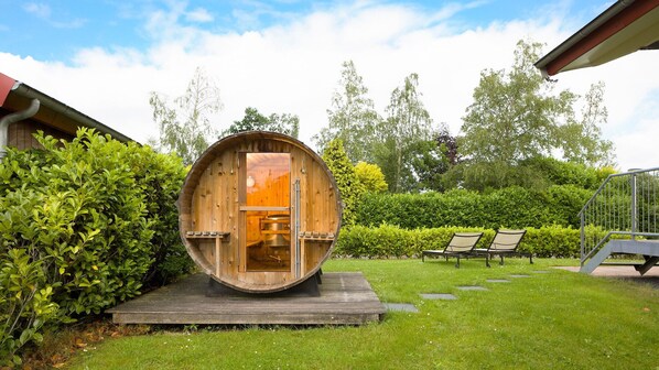 A wooden barrel structure with a door, surrounded by a wooden deck, and two lounge chairs in a garden setting.