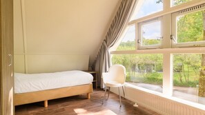 Un dormitorio con una cama individual, una silla y ventanas amplias que dan a vistas de vegetación y una casa.