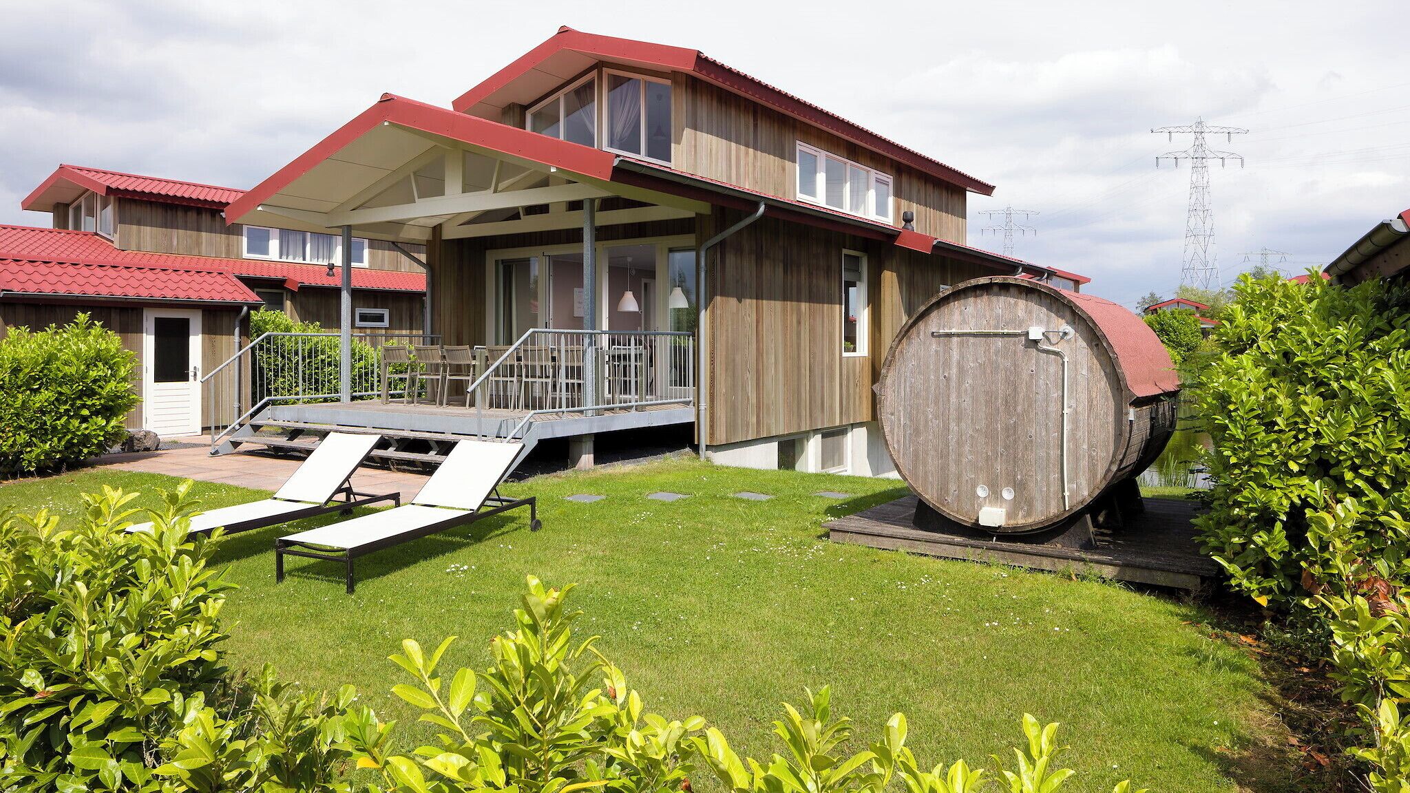 Een modern gebouw met een houten gevel, een overdekt terras met stoelen en een groot vat op de voorgrond.