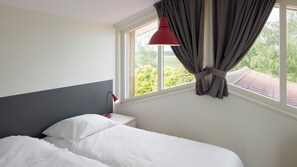 A hotel room with a bed, a nightstand, a red lamp, and a window with curtains.