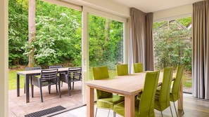 Un comedor con una mesa de madera y sillas verdes, ventanas amplias con cortinas y vistas a un jardín exuberante.