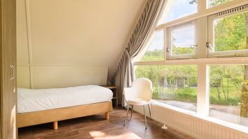 A bedroom with a single bed, a chair, and large windows offering a view of greenery and a house.