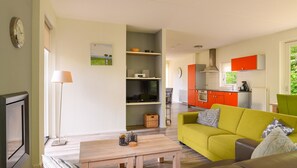 Una sala moderna con sofá, una mesa de centro y televisor. Se observa una cocina con armarios naranjas y una ventana.