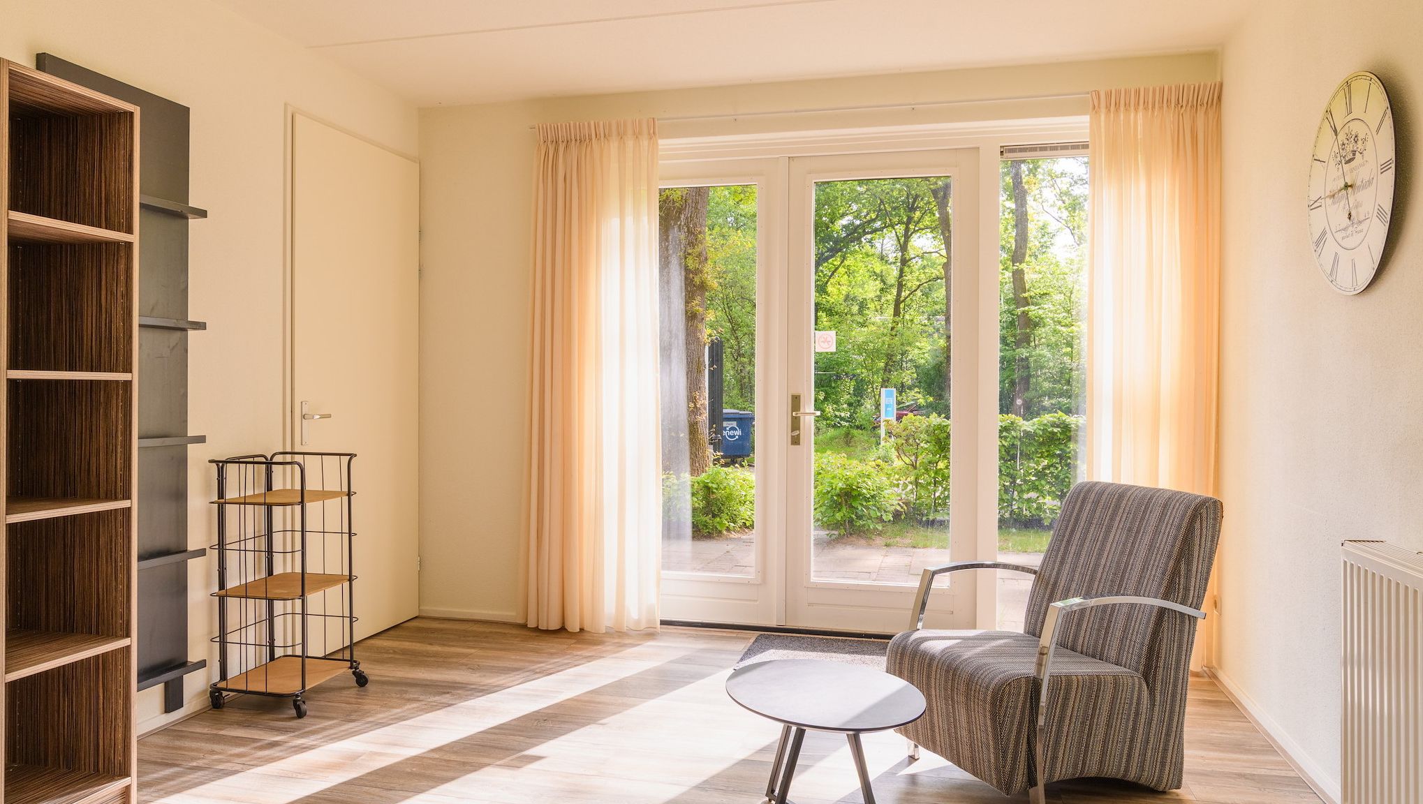 A room with a sliding glass door leading to a garden, a chair, a small table, and a bookshelf.