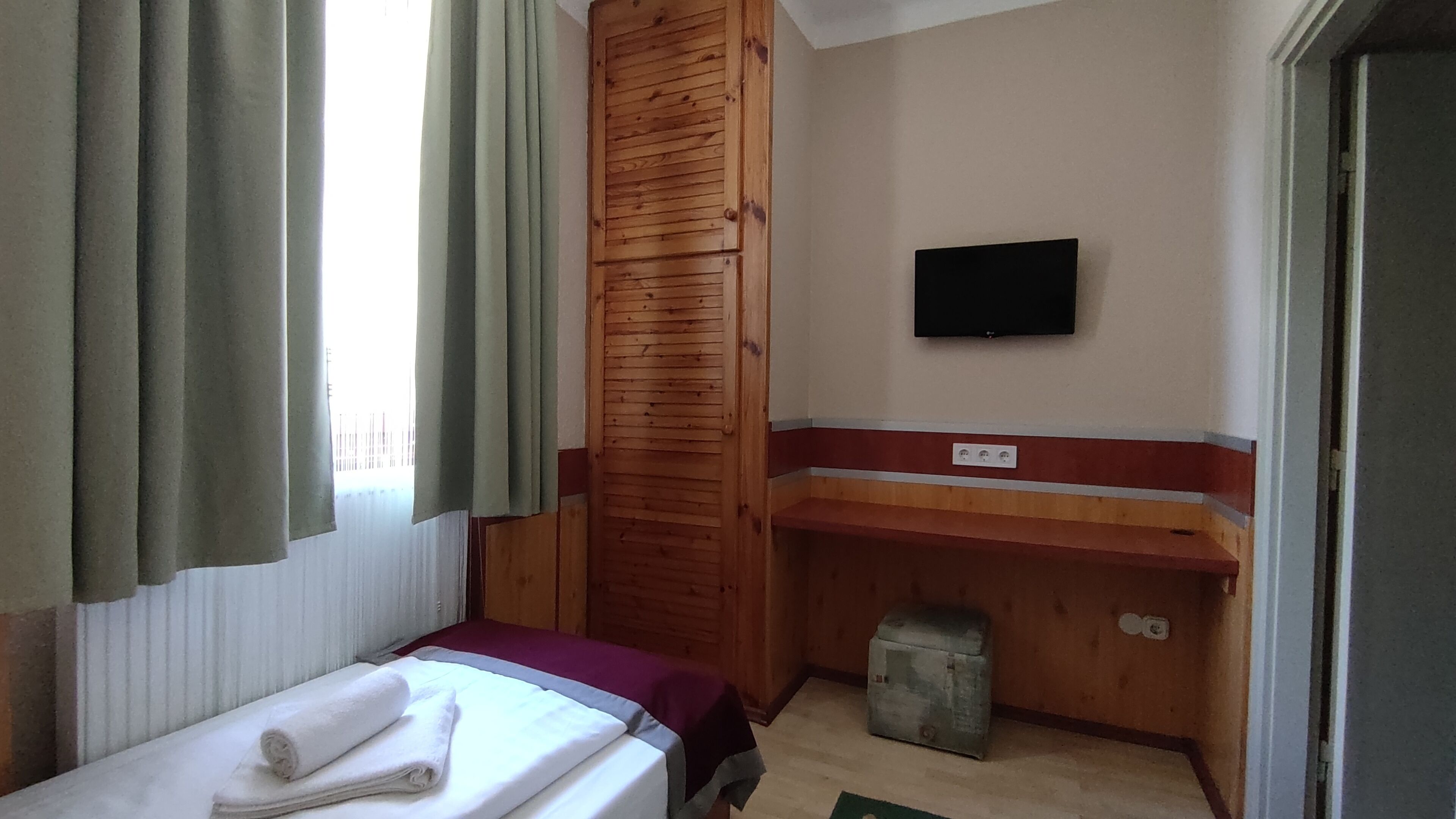 Una habitación de hotel pequeña con una cama, un armario de madera, un televisor fijado a la pared y un pequeño cubo de basura.