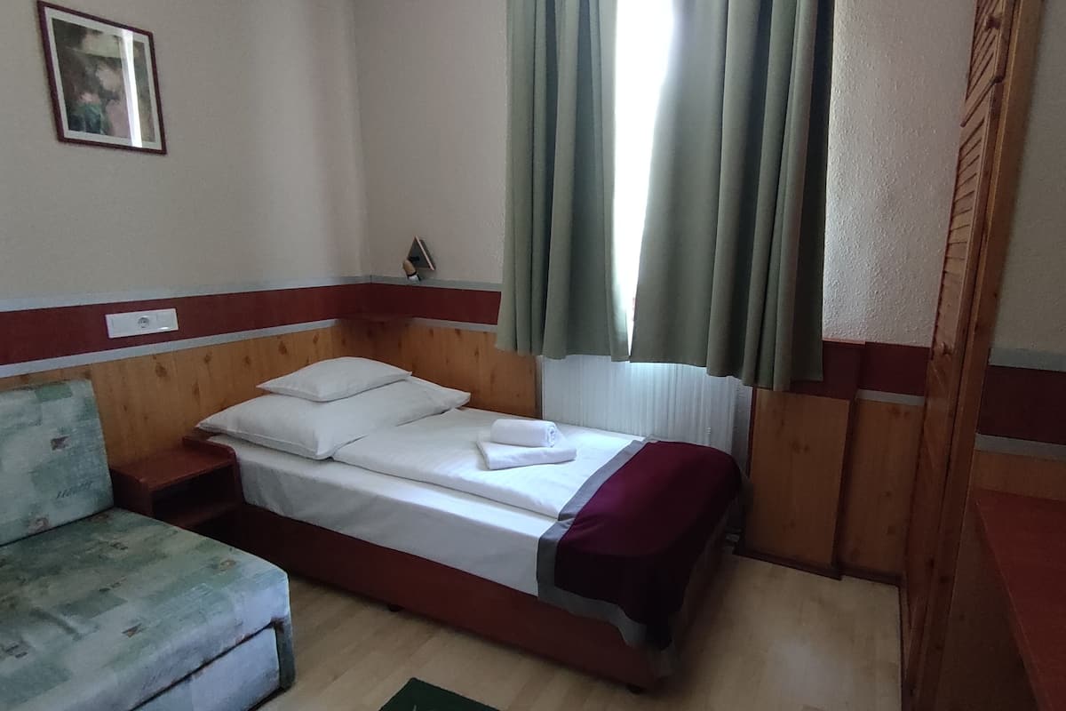Una cama individual con sábanas blancas y una colcha roja, un cabecero de madera, una mesita de noche pequeña y una cortina verde.