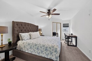 Ein Schlafzimmer mit einer gepolsterten Kopfteil, einem Bett mit floraler Bettwäsche, einem Schreibtisch, einem Stuhl, einem Deckenventilator und einem Fenster mit Jalousien.