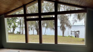Un patio cubierto con mobiliario de mimbre y un ventanal grande con vista a un lago rodeado de árboles y un muelle.