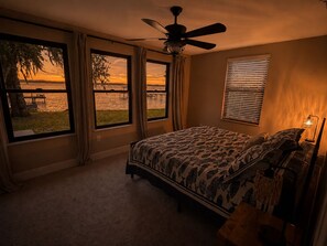 Une chambre à coucher avec un lit, un ventilateur de plafond, une fenêtre donnant sur un coucher de soleil et une lampe.