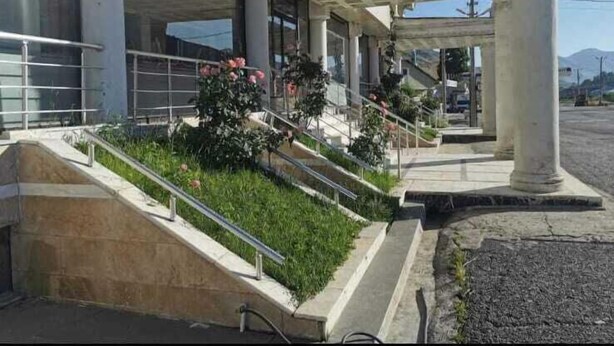 A building with a covered entrance, steps leading up to it, and a garden area with plants and flowers.